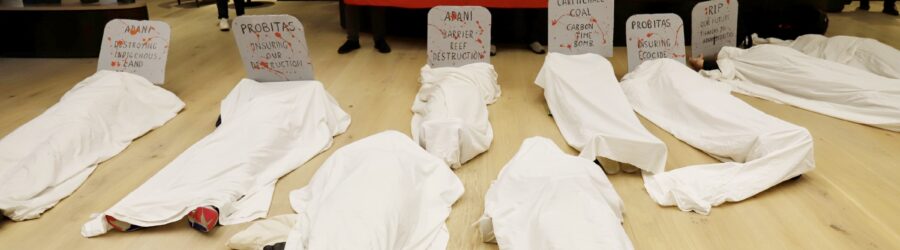 Bodies Lay On The Floor Covered In White Sheets For The "die In" Action. People Stand Behind With A Red Sign That Says "Probitas Stop Insuring Adani"