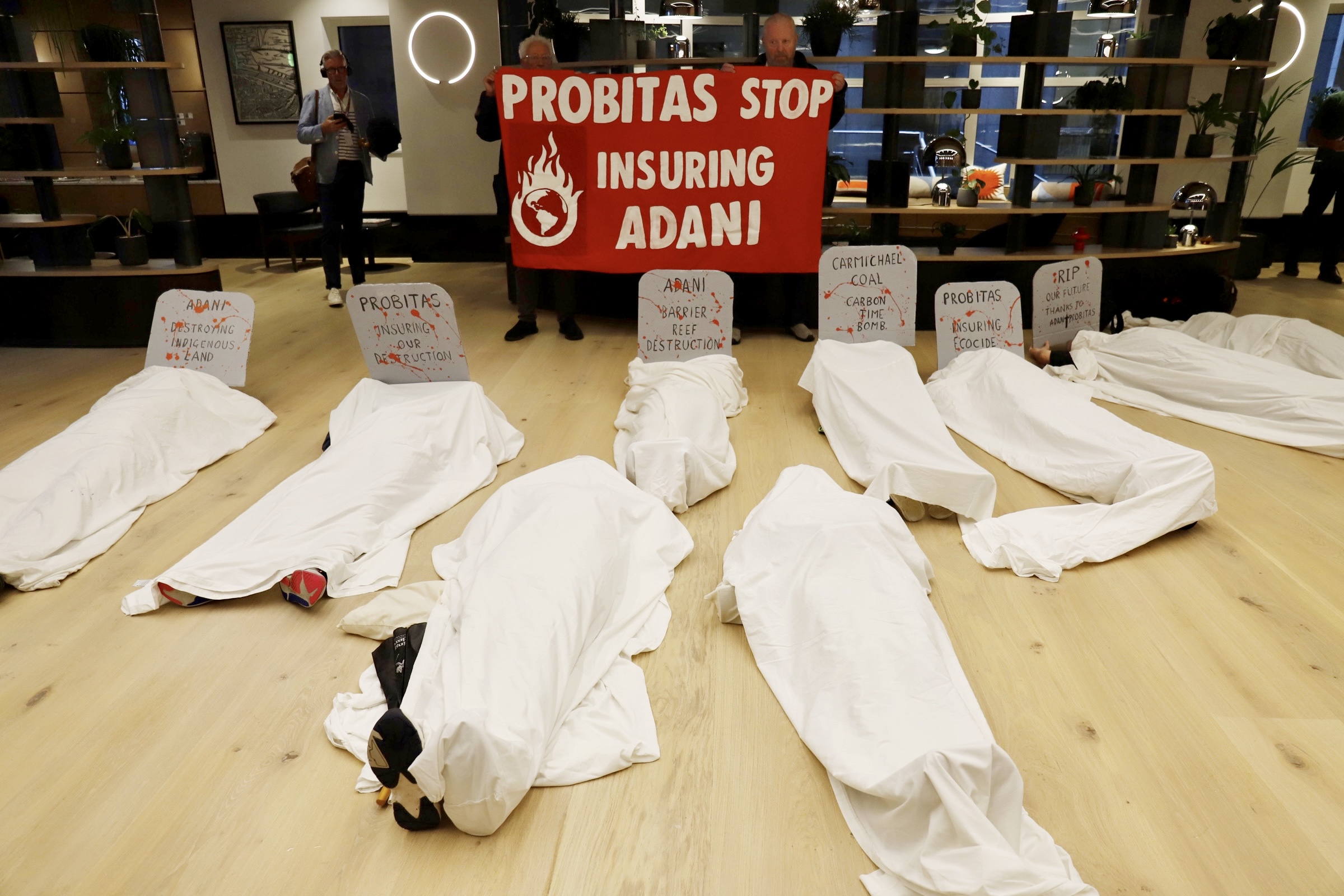 Bodies lay on the floor covered in white sheets for the "die in" action. People stand behind with a red sign that says "Probitas Stop Insuring Adani"